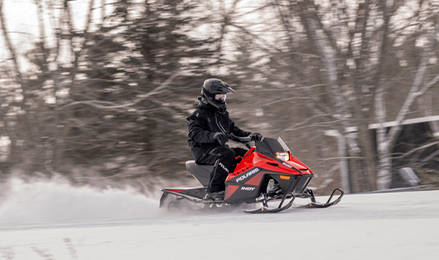 A rider speeds across a snow-covered trail on a red Polaris INDY 200 snowmobile with a blurred winter landscape background.