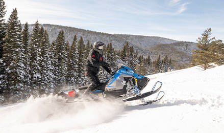 A rider on a blue and grey Polaris snowmobile Voyageur Nordic Pro with the 650 Patriot engine, navigates a snowy hillside with a forest and mountains in the distance.