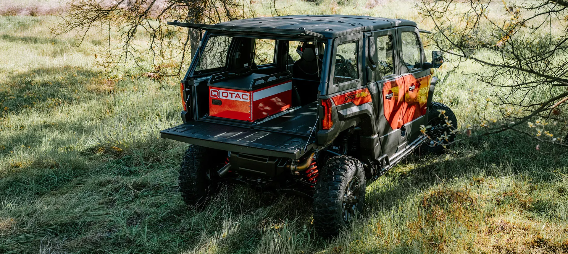 Polaris XPEDITION ADV 5 NorthStar off-road vehicle in grassy terrain with rear cargo bed open, carrying a mounted QTAC unit beneath tree cover in a rural landscape.