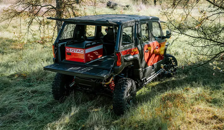 Polaris XPEDITION ADV 5 NorthStar off-road vehicle in grassy terrain with rear cargo bed open, carrying a mounted QTAC unit beneath tree cover in a rural landscape.