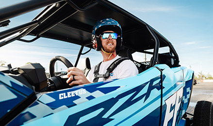 Cleetus McFarland wearing a stars-and-stripes helmet and blue-mirrored sunglasses, sits at the wheel of a custom blue and white Polaris RZR Pro R 4 Ultimate with CLEETUS and FF (Freedom Factory) decals.
