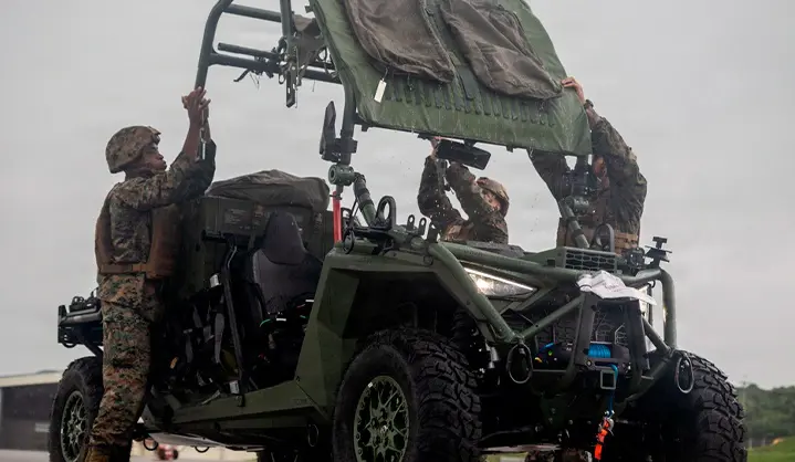 Military personnel loading equipment onto a Polaris MRZR military Side-by-Side vehicle in wet outdoor conditions with tactical gear mounted throughout the vehicle.