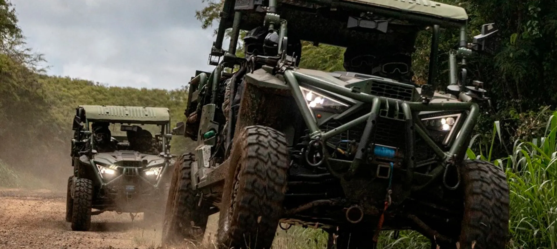 Two Polaris MRZR military Side-by-Side vehicles driving along a muddy jungle trail with headlights illuminated and tactical equipment mounted on the frames.