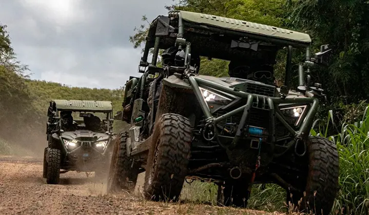 Two Polaris MRZR military Side-by-Side vehicles driving along a muddy jungle trail with headlights illuminated and tactical equipment mounted on the frames.
