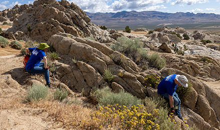 Volunteers in blue Polaris shirts collecting debris and restoring vegetation among large desert rock formations, with mountains and dry valley terrain in the background.