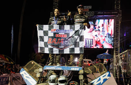 Off-road racing team standing on a Polaris RZR race vehicle while holding a BFGoodrich SCORE Baja 500 banner on a nighttime podium stage with event screens and lighting in the background.