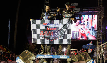 Off-road racing team standing on a Polaris RZR race vehicle while holding a BFGoodrich SCORE Baja 500 banner on a nighttime podium stage with event screens and lighting in the background.