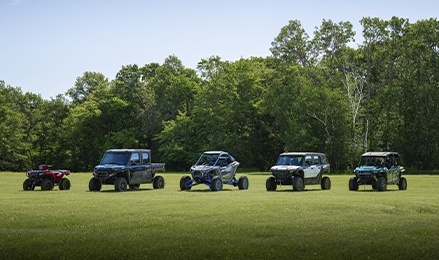 Polaris Sportsman ATV, RANGER UTV, RZR and XPEDITION Side-by-Sides lined up on a grassy field with a wooded backdrop.