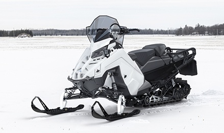 White Polaris Military 850 Voyageur 155 Crossover snowmobile with a windshield, two-up seat and rear cargo rack parked in a snowy field.