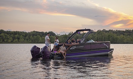 Godfrey AquaPatio luxury pontoon boat with twin outboard engines carrying passengers and a dog on a lake at sunset.