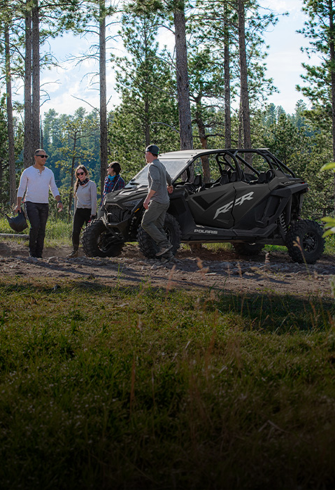 Polaris RZR Pro XP 4 Side-by-Side parked in a forest, with four people standing beside it among tall pine trees in a grassy wooded setting.