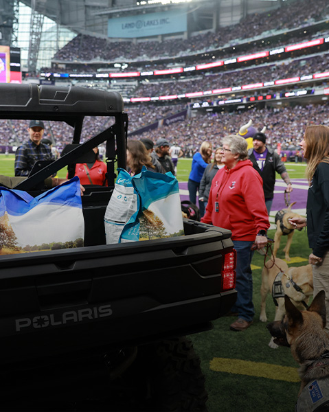 Polaris RANGER UTV parked on a stadium field with its cargo bed loaded with large bags, surrounded by people and service dogs during an event.
