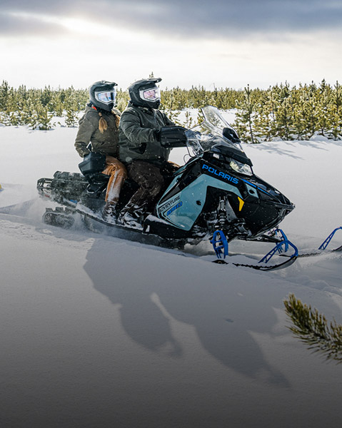 Polaris Voyageur 650 snowmobile towing a cargo sled through a snowy forest landscape, with two riders wearing helmets as the machine moves across fresh snow.