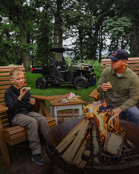 Polaris RANGER 500 UTV parked on grass in a wooded area, with a father and his son seated in wooden chairs in the foreground sharing food near a small campfire.
