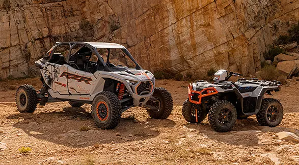 A white and grey Polaris RZR Pro R 4 SxS and a Polaris Sportsman XP 1000 ATV are parked side-by-side in a rocky desert landscape.