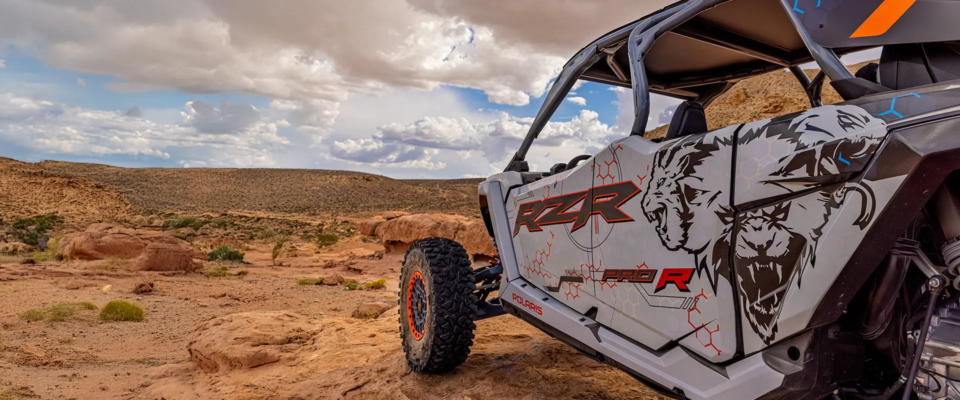 A white and grey Polaris RZR Pro R 4 SxS is parked on a rocky outcrop with a vast desert landscape and cloudy sky in the background.