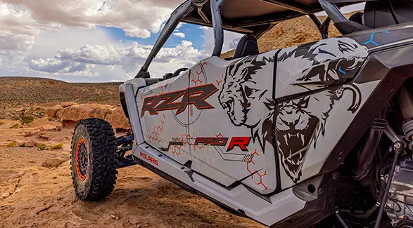 A white and grey Polaris RZR Pro R 4 SxS is parked on a rocky outcrop with a vast desert landscape and cloudy sky in the background.