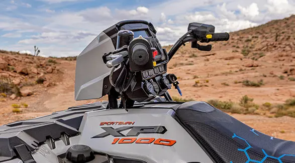 A close-up of the handlebars and front rack of a white and grey Polaris Sportsman XP 1000 ATV in a desert setting.