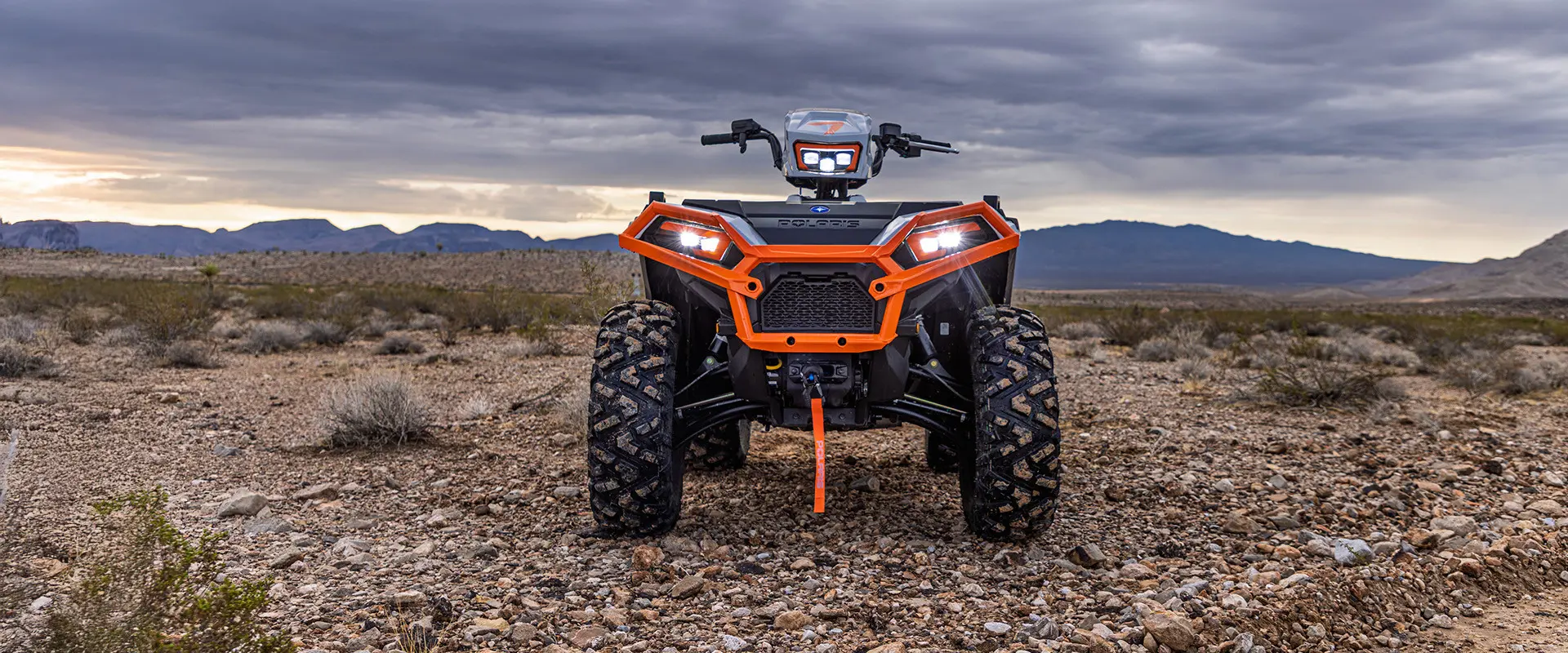 A front view of an orange and grey Polaris Sportsman XP 1000 ATV parked in a desert landscape under a dramatic sky.