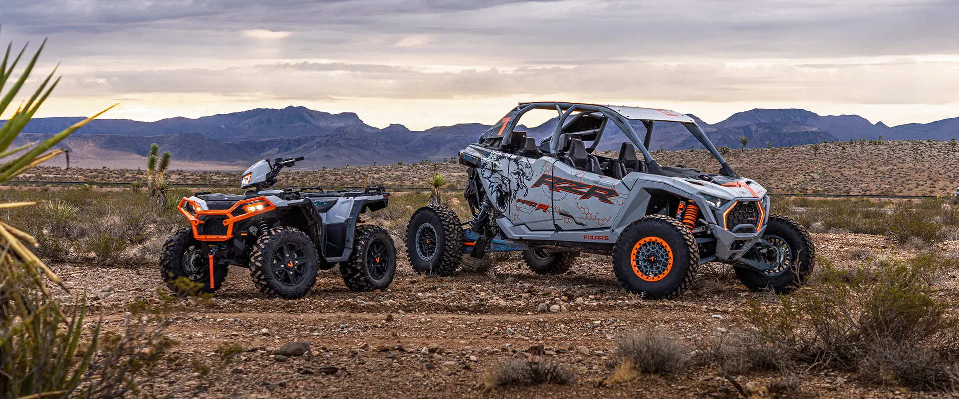 A white and grey Polaris RZR Pro R 4 SxS and a Polaris Sportsman XP 1000 ATV are parked in a desert with mountains in the background under an overcast sky.