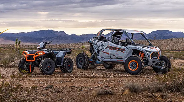 A white and grey Polaris RZR Pro R 4 SxS and a Polaris Sportsman XP 1000 ATV are parked in a desert with mountains in the background under an overcast sky.