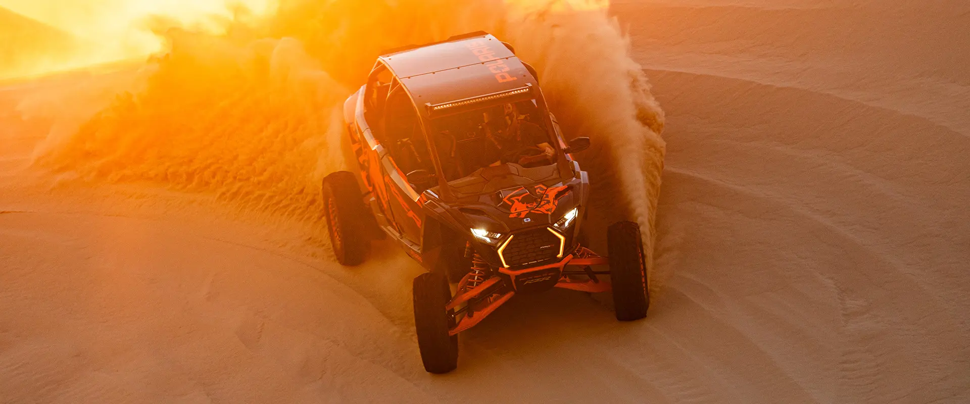 Un VHR côte à côte Polaris RZR Pro R 2025 orange et noir soulève un grand jet de sable alors qu’il traverse une dune désertique.