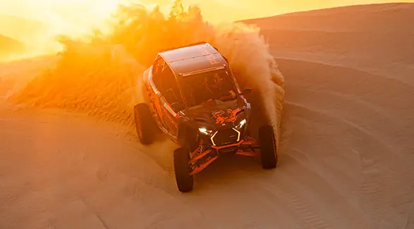 Un VHR côte à côte Polaris RZR Pro R 2025 orange et noir soulève un grand jet de sable alors qu’il traverse une dune désertique.