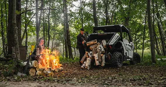 Deux personnes près d’un véhicule GENERAL XP 1000 qui décharge du bois de chauffage à côté d’un feu de camp