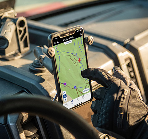 A gloved hand interacting with the Polaris RIDE COMMAND app on a smartphone mounted inside a Polaris Off Road vehicle, displaying a detailed trail map.