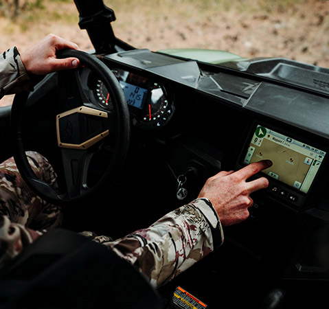 A driver in camouflage clothing navigates a Polaris GENERAL UTV, interacting with the RIDE COMMAND touchscreen display showing a trail map.