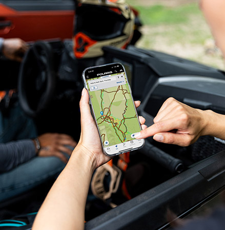 A person standing outside a Polaris GENERAL UTV holds a smartphone displaying the Polaris RIDE COMMAND trail map, pointing at the screen while a driver sits inside the vehicle.