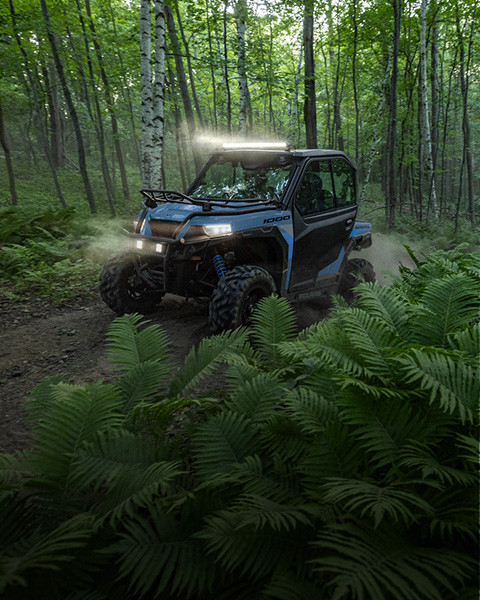 Blue Polaris GENERAL 1000 UTV driving through a lush green forest with dust trailing, lights on.