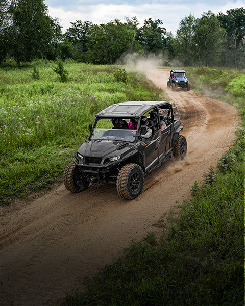 Black Polaris GENERAL XP 1000 UTV with four passengers driving on a dirt road, kicking up dust amidst green foliage.