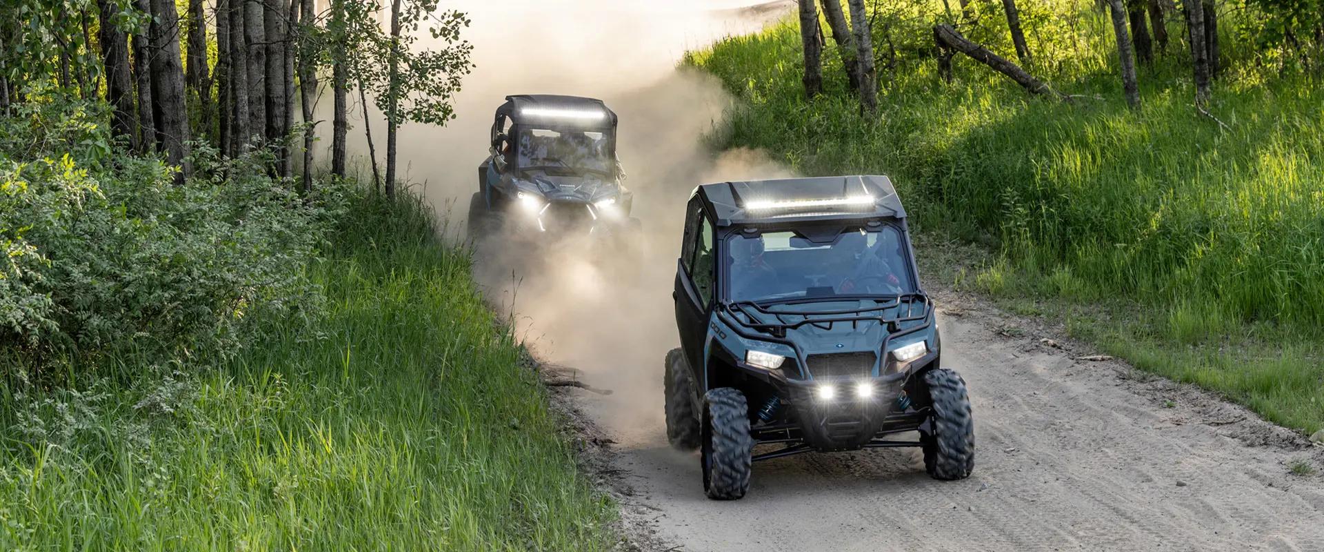 Two 2026 Polaris GENERAL crossover side-by-sides cruising on a dusty trail with illuminated headlights.