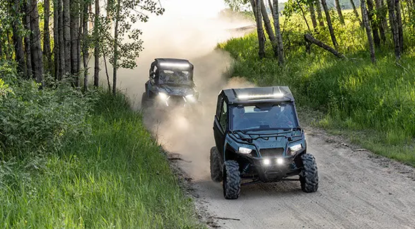 Two 2026 Polaris GENERAL crossover side-by-sides cruising on a dusty trail with illuminated headlights.