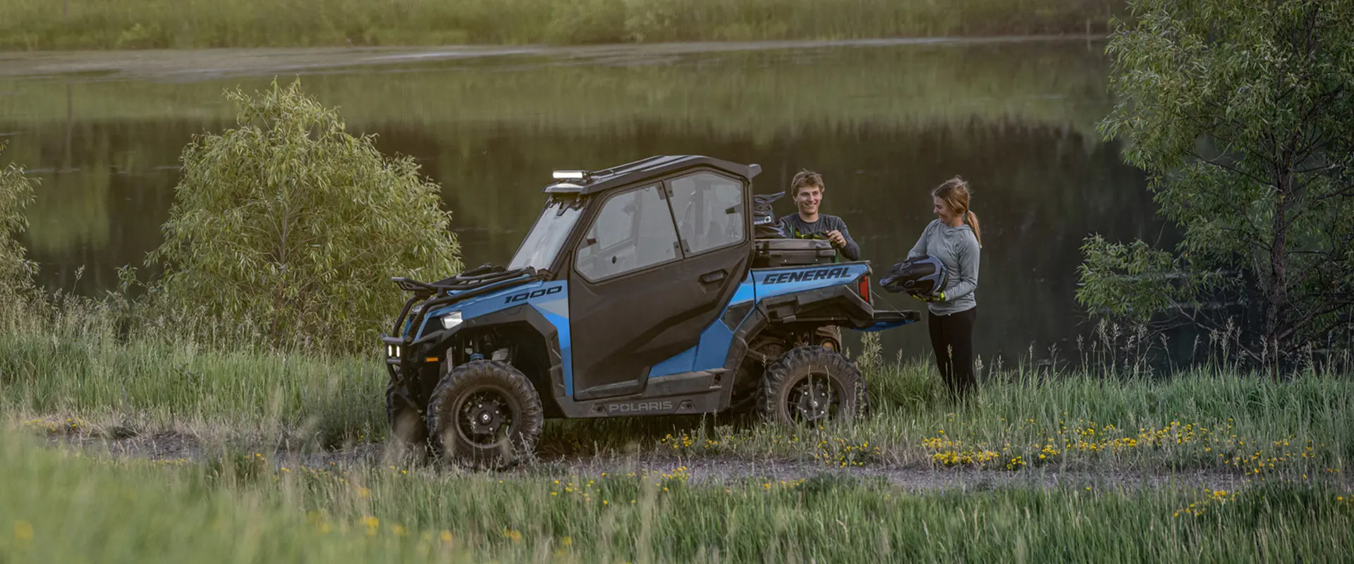 A couple standing by a lake and retrieving items from the cargo box of their 2026 Polaris GENERAL crossover side-by-side.