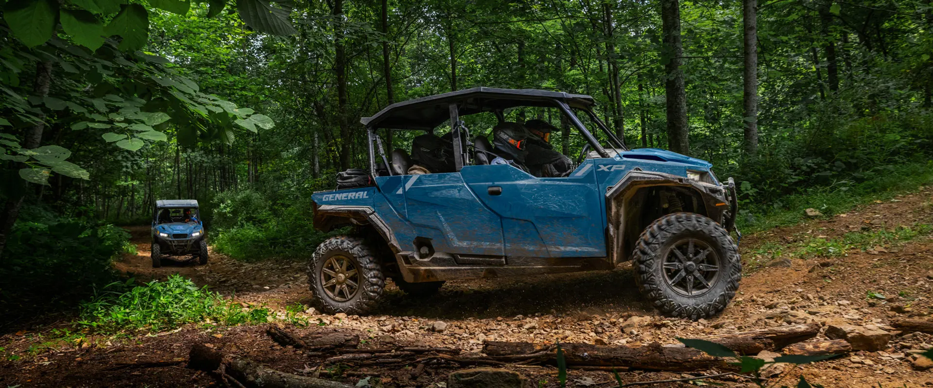 Side profile of a 2026 Polaris GENERAL XP 4 1000 crossover side-by-side cruising through the forest while another GENERAL trails behind.