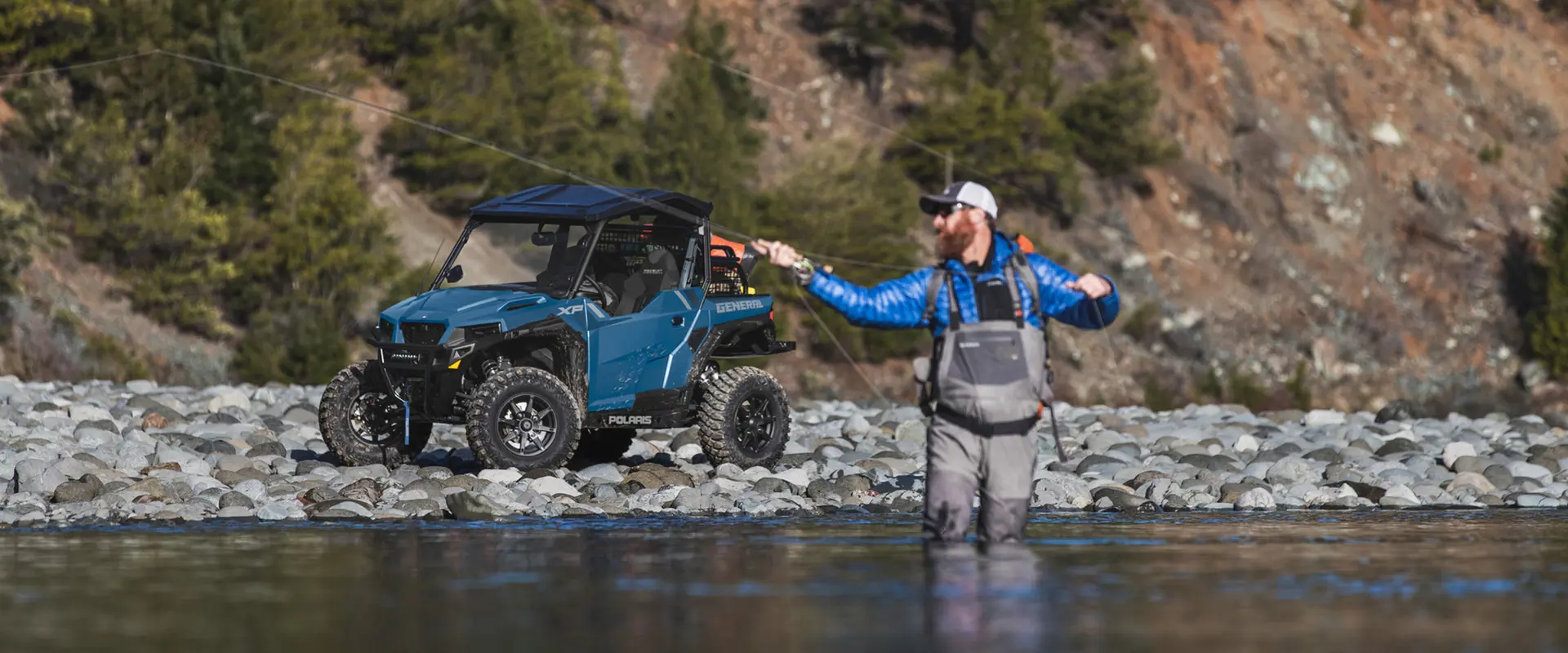 A man fishing in a river while his 2026 Polaris GENERAL XP 1000 crossover side-by-side is parked in the background.