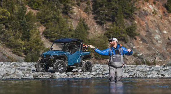 A man fishing in a river while his 2026 Polaris GENERAL XP 1000 crossover side-by-side is parked in the background.