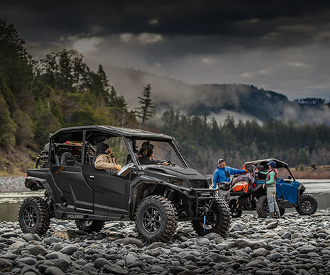Polaris GENERAL UTVs equipped with off-road accessories including roof racks, storage bags, and rugged tires, parked on rocky terrain near water with outdoor enthusiasts.