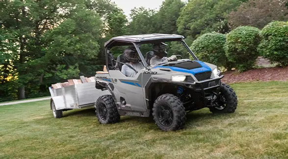 2 riders in a 2027 Polaris GENERAL 1000 side-by-side pulling a loaded trailer across the lawn.