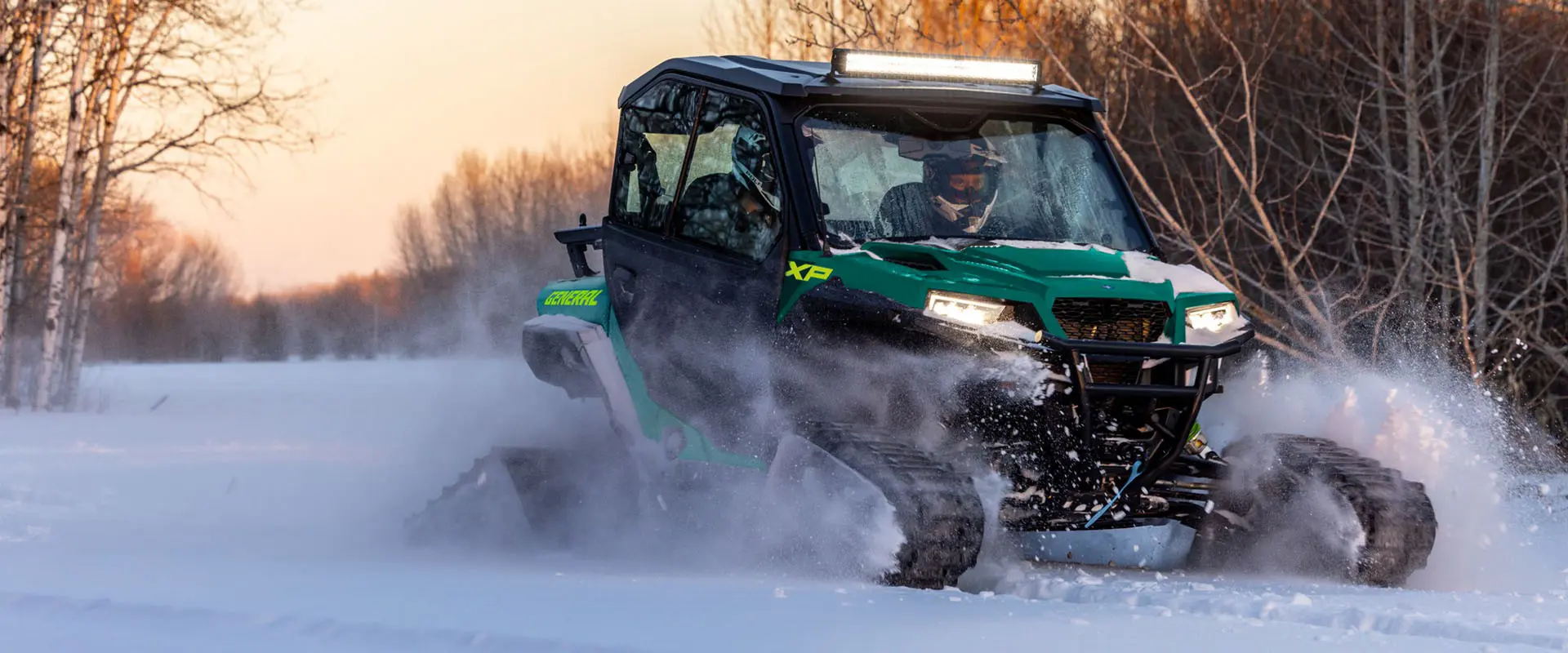 2 riders in a 2027 Polaris GENERAL XP 1000 side-by-side with snow tracks installed driving through a snowy field.