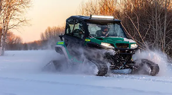 2 riders in a 2027 Polaris GENERAL XP 1000 side-by-side with snow tracks installed driving through a snowy field.
