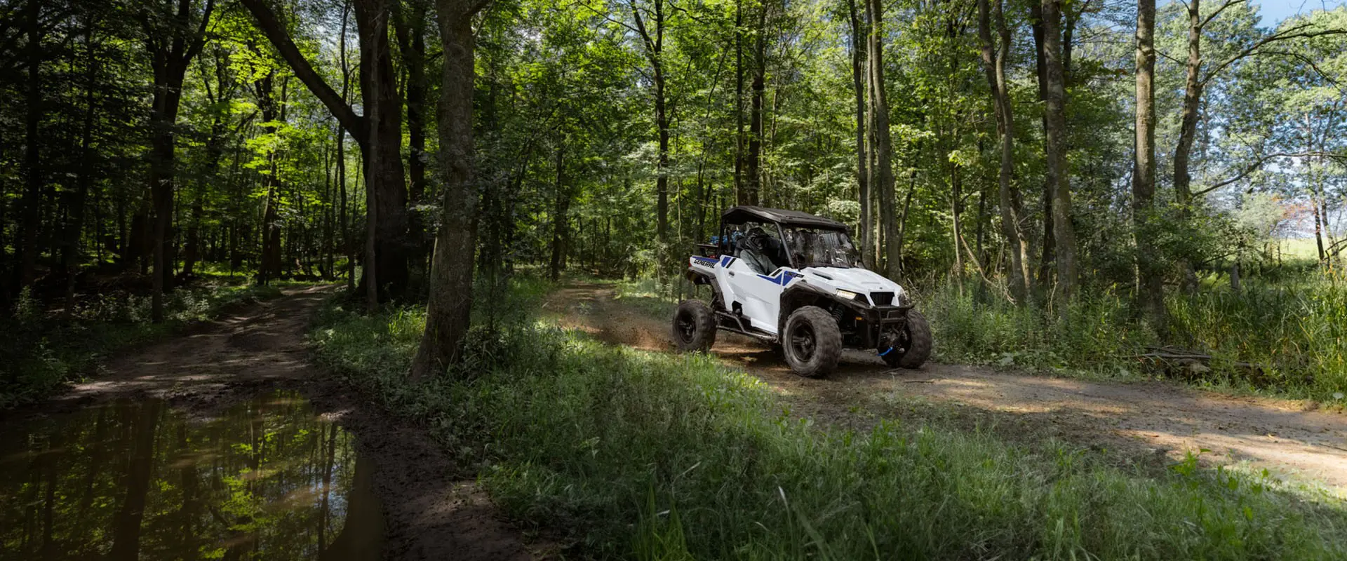 2 riders in a 2027 Polaris GENERAL XP 1000 side by side enjoying a forested trail ride on a beautiful day.