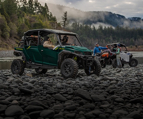 Polaris GENERAL UTVs equipped with off-road accessories including roof racks, storage bags, and rugged tires, parked on rocky terrain near water with outdoor enthusiasts.