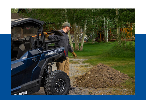 A man behind his General, pulling out a shovel from the Rhino rack accessory. looking at a pile of dirt