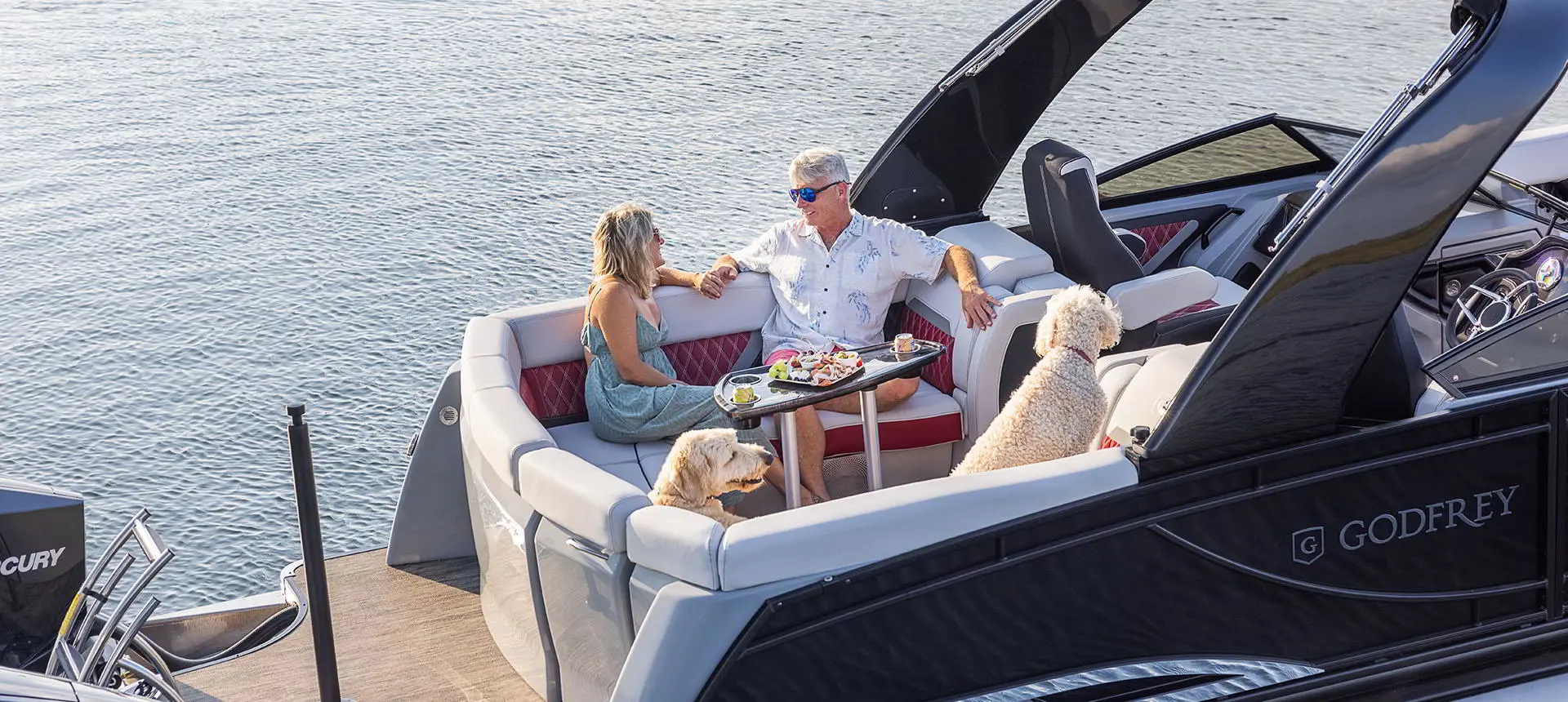 Couple prenant une collation avec leurs chiens à la table du vaste coin salon à la poupe d’un ponton Godfrey Sanpan Lounge 2700 ULW Twin.