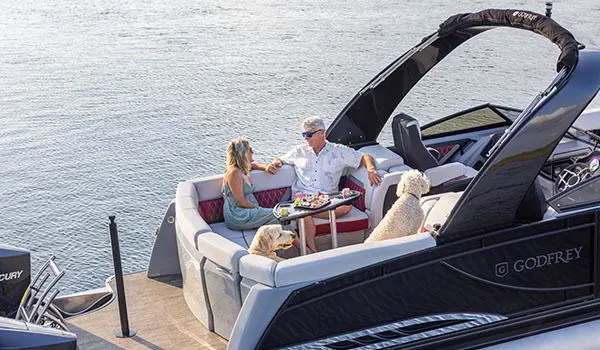 Couple prenant une collation avec leurs chiens à la table du vaste coin salon à la poupe d’un ponton Godfrey Sanpan Lounge 2700 ULW Twin.