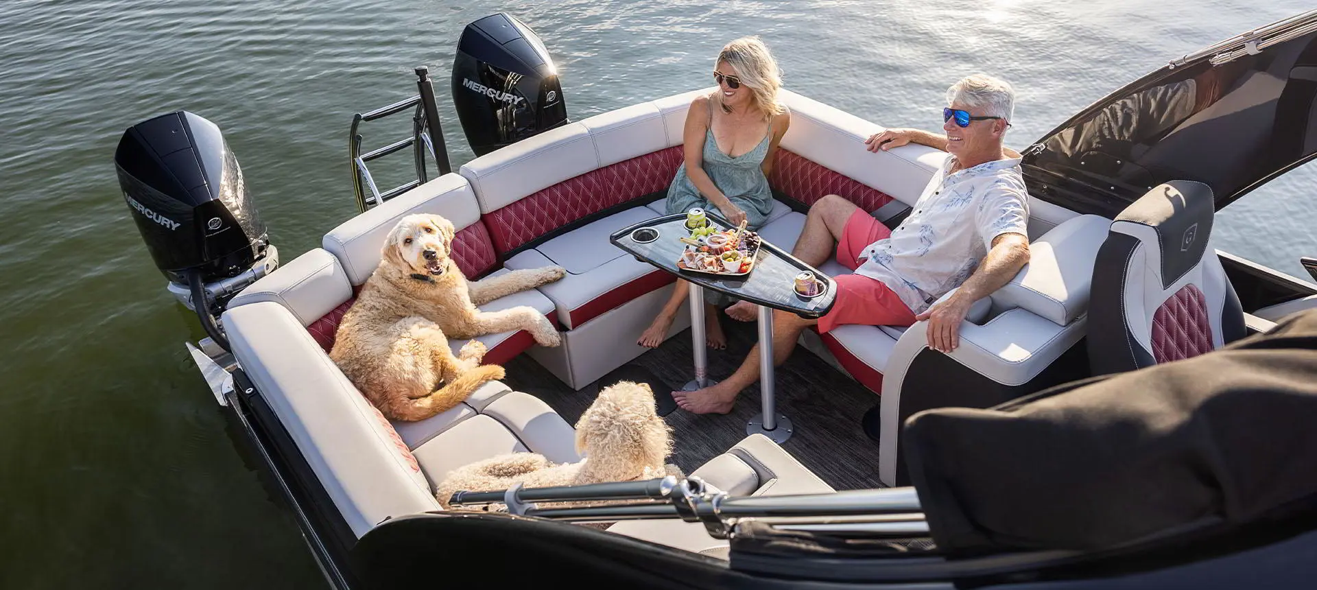 Vue aérienne d’un couple prenant une collation avec leurs chiens dans le vaste coin salon à la poupe d’un ponton Godfrey Sanpan Lounge 2700 ULW Twin.