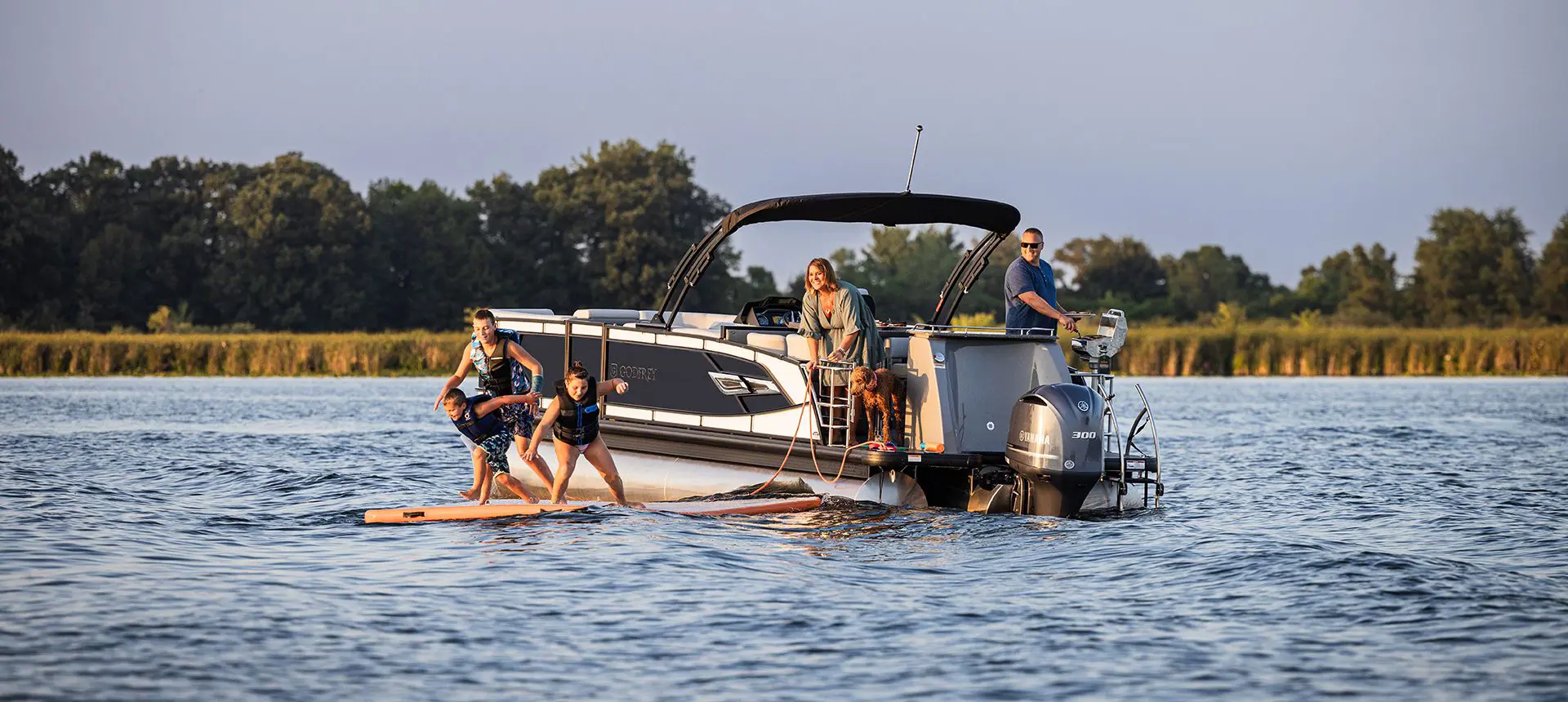 Kids playing on a stand-up paddle board near a Godfrey AquaPatio Entertainment pontoon boat on a lake.